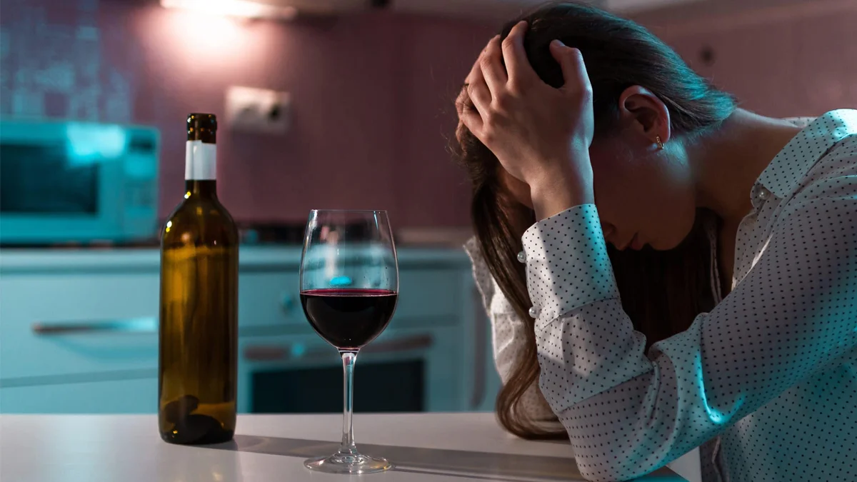 Woman sitting with a glass of red wine in the dark with her head in her hands. Alcohol is classified as a depressant drug